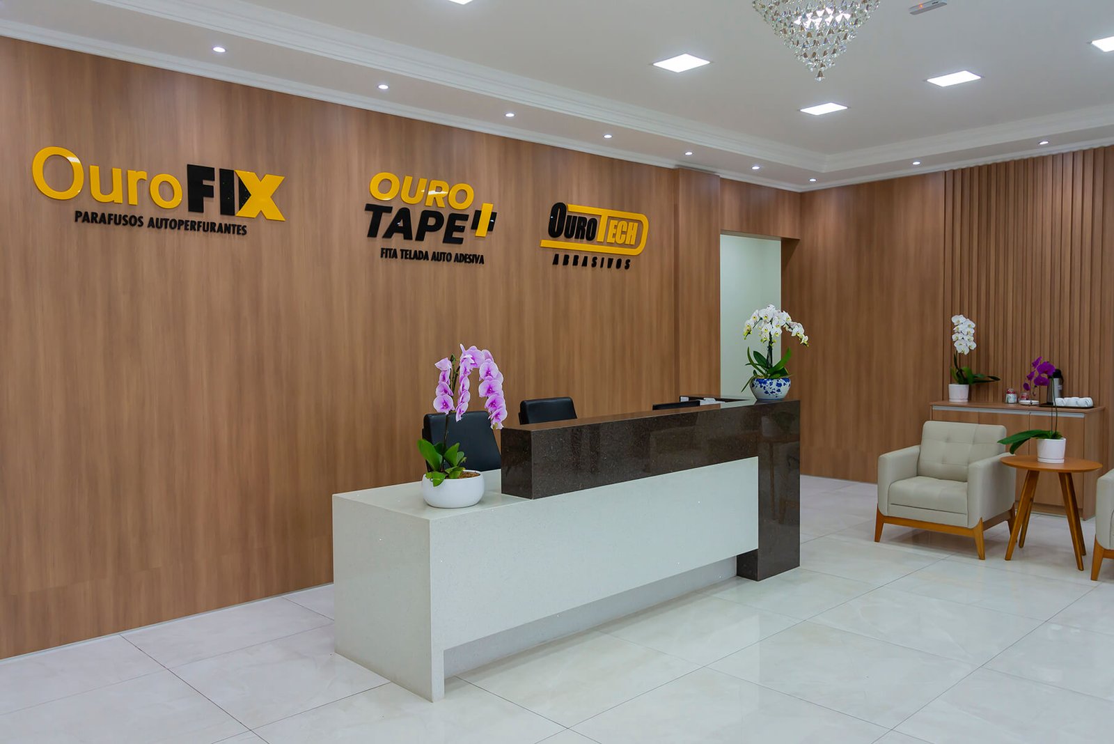 Em nossa recepção temos um balcão de atendimento branco e preto. Atrás deste balcão temos as logos da Ourofix, Ourotape e ourotech, 3 marcas comercializadas em nosso catálogo.